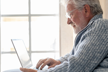 Elderly 70s man seated on sofa browsing on laptop, senior looking at laptop screen surfing the net,...