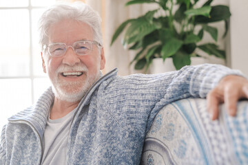 Portrait of happy senior man wearing eyeglasses smiling looking at camera. Old man relaxing on sofa at home. Portrait of elderly man enjoying retirement.