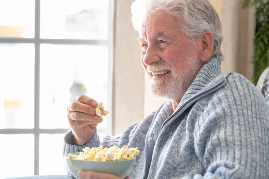 Senior 70s man seated on sofa eating popcorn, leisure and people concept - happy bearded senior man relaxing at home looking at tv