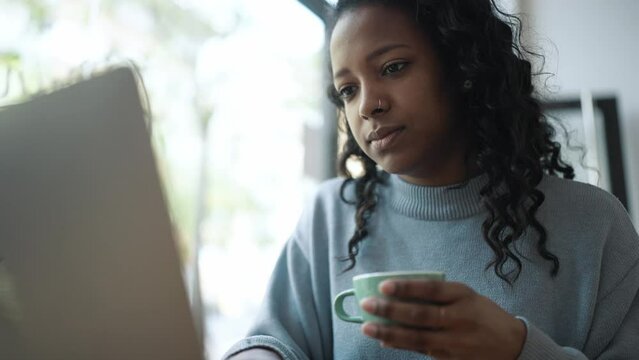 Confident African woman wearing blue sweater typing by laptop with cup of coffee in hands in cafe