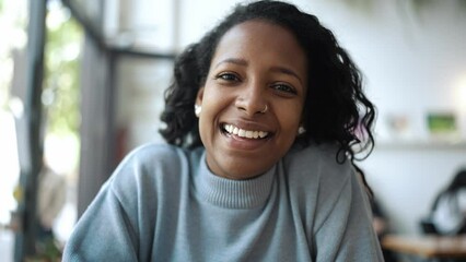 Smiling African woman wearing blue sweater talking by video call on laptop and making greeting gesture in cafe