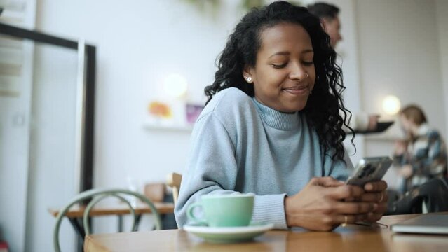 Happy African woman wearing blue sweater texting on phone and looking to the side in cafe