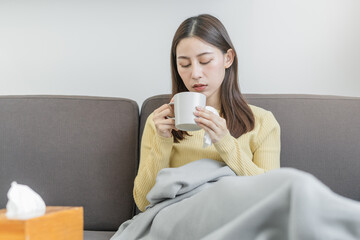 Sick, hurt or pain asian young woman, girl sore throat with glass, mug of warm water, headache have a fever, flu in weakness, sitting relaxed on sofa bed at home. Health care person on virus seasonal.