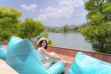 Beautiful smiling young woman wearing hat sitting on beach bean bag furniture Blue color chair on wooden balcony with lake view and sky background in summer holiday vacation. Leisure, travel, tourism.