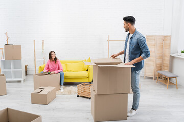 Cheerful woman holding book near arabian boyfriend and packages at home.