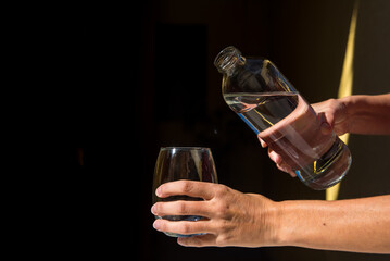 delicate hands of woman holding glass bottle with water and a glass tumbler, black background, concept of vitality, health, wellbeing