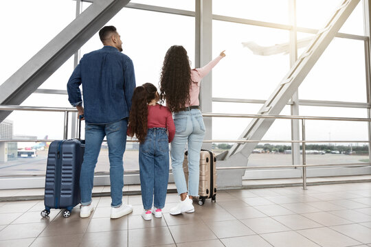 Family Of Three Looking Out Of Window At Airport And Pointing Away