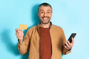 Middle Aged Man Holding Cellphone Showing Credit Card, Blue Background