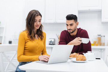 Smiling woman using laptop near muslim man and breakfast in kitchen.