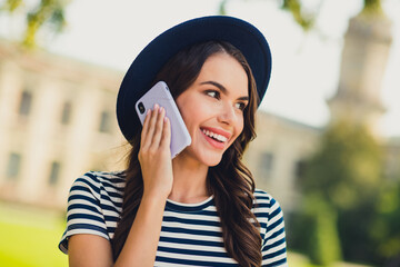 Photo of sweet pretty young lady wear striped dress headwear smiling walking talking modern device outdoors urban city park