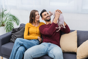 Positive multiethnic couple looking at sphynx cat on couch.
