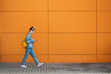 Cute teenage girl with a gadget and headphones. Against the background of an orange wall. The concept of modern youth. Kids with gadgets. Modern world. Lifestyle. Minimalism.