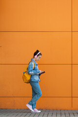 Fototapeta na wymiar Cute teenage girl with a gadget and headphones. Against the background of an orange wall. The concept of modern youth. Kids with gadgets. Modern world. Lifestyle. Minimalism.