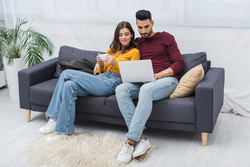 Obraz premium Smiling woman holding cup near muslim boyfriend with laptop on couch at home.