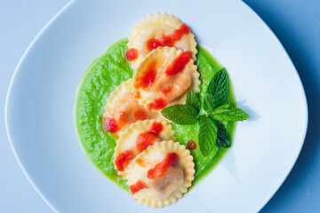 Italian pasta ravioli with burrata filling with tomato and pea cream on a white plate shot from above with a light blue background in horizontal shot