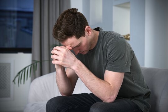 Sad Upset Depressed Unhealthy Guy, Young Tired Exhausted Frustrated Man Is Suffering, Holding His Head With Hand At Home, Sitting On Couch Or Sofa, Having Headache, Migraine. Failure, Despair Concept