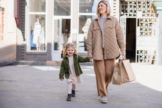 trendy woman with shopping bags and cheerful girl holding hands while walking on urban street.