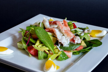 salad with crab meat and tomatoes with avocado and greens with poached egg on white plate on dark table