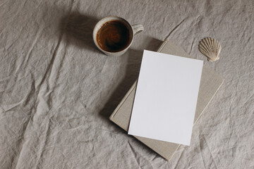 Feminine stationery, desktop mock-up scene. Blank greeting card, craft envelope, cup of coffee, sea shell and old book on beige linen table background. Flat lay, top view. Summer moody composition.