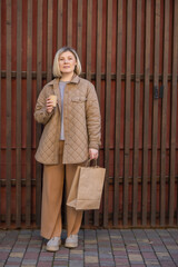 full length view of trendy woman with coffee to go and shopping bag looking at camera near wooden fence.