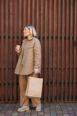 full length view of stylish woman standing with paper cup and shopping bag near wooden fence.