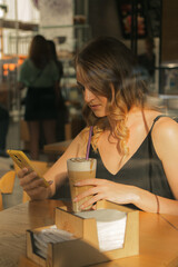 A pretty young woman drinks coffee in a cafe and chats on a smartphone, view through glass