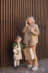 happy woman with paper cup talking on smartphone near daughter with teddy bear and shopping bag.