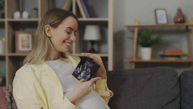Expectant caucasian mother holding ultrasound scan while sitting on comfy sofa in living room. Pregnant woman enjoying future motherhood with first sonogram image of unborn baby.