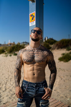 Chico joven rapado con tatuajes y musculado posando con ropa actual en la calle con mucho sol