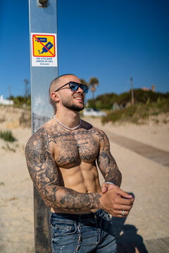 Chico joven rapado con tatuajes y musculado posando con ropa actual en la calle con mucho sol