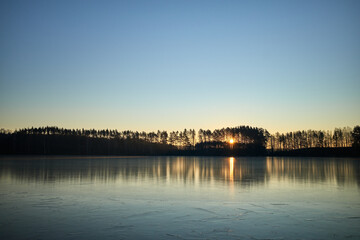 Fototapeta premium Sunrise behind the trees is reflected on the ice surface of the