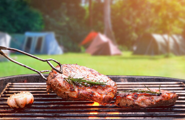 Roasted beef ripe steaks on flaming charcoal grill with blurred background of camping area in...