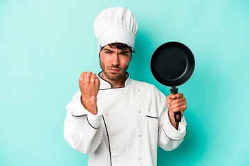 Young caucasian chef man holding flying pan isolated on blue background showing fist to camera,...