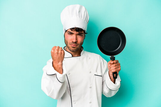Young caucasian chef man holding flying pan isolated on blue background showing fist to camera, aggressive facial expression.