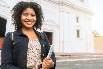Curly-haired mestizo university student with a backpack and a notebook in her hand smiling and looking at the camera outdoors in the central park of leon