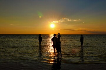 A small group of silhouetted young people wade in shallow waters and watch the sunset on a summer evening at Rock Harbor on Cape Cod.