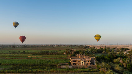 Bright balloons are flying over the Nile Valley. Cultivated green fields, palm trees, houses are visible. In the distance - sand dunes. Clear blue sky. Egypt. Luxor