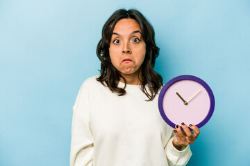 Young hispanic woman holding a clock isolated on blue background shrugs shoulders and open eyes...