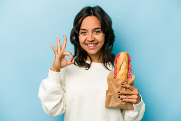 Young hispanic woman eating a sandwich isolates on blue background cheerful and confident showing...