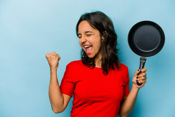 Young hispanic woman holding a frying pan isolated on blue background raising fist after a victory,...