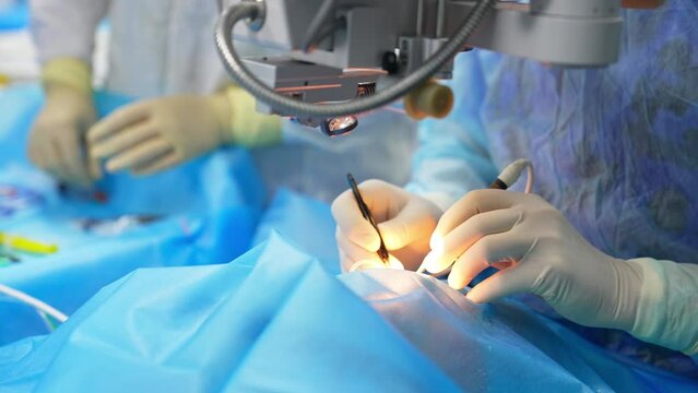 Surgeon's hands in latex gloves apply instruments in the patient's eye. Equipment over the table sending light to the operated area. Close up.