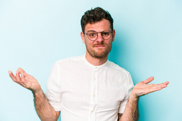 Young caucasian man isolated on blue background doubting and shrugging shoulders in questioning...