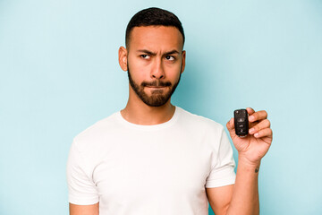 Young hispanic man holding car keys isolated on blue background confused, feels doubtful and unsure.
