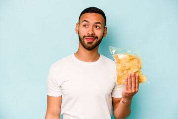 Young hispanic man holding a bag of chips isolated on blue background dreaming of achieving goals...
