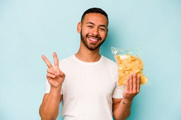 Young hispanic man holding a bag of chips isolated on blue background joyful and carefree showing a...