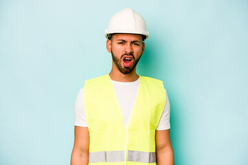 Young laborer hispanic man isolated on blue background screaming very angry and aggressive.