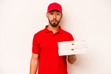 Young hispanic delivery man holding pizzas isolated on white background shrugs shoulders and open...