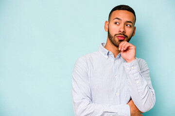 Young hispanic man isolated on blue background looking sideways with doubtful and skeptical expression. - obrazy, fototapety, plakaty