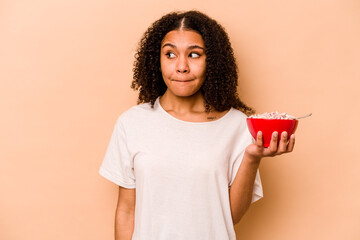 Young African American woman holding a bowl of cereals isolated on beige background confused, feels doubtful and unsure.