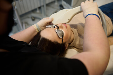 Young pretty woman wearing UV protective goggles, lying down on daybed while receiving hair removal laser procedure on her face in a wellness spa clinic